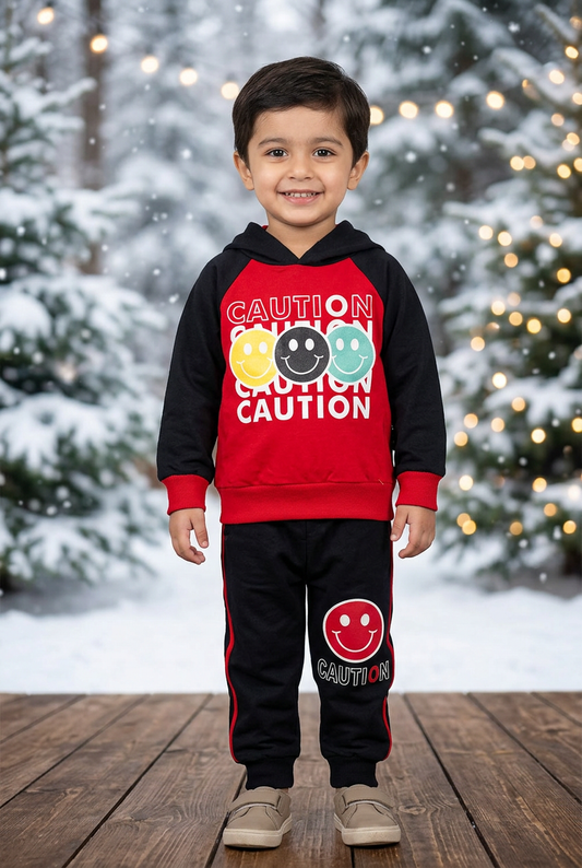 Child wearing BS827 red and black Caution smiley Terry hoodie and black red-striped joggers on a sheltered veranda beside snowy pines and warm lights.